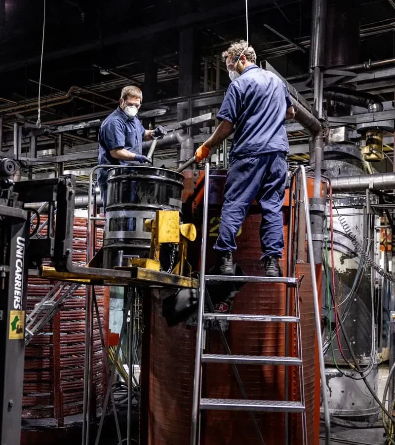 Two FMI employees in full protective gear standing on either side of a metal drum and stirring the contents inside.